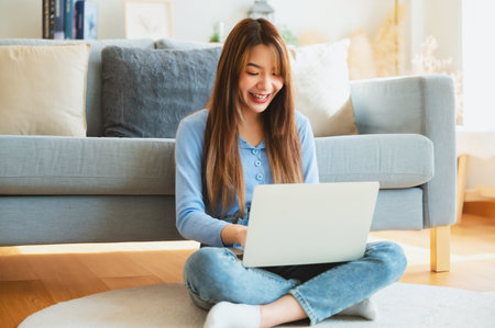 Young asian woman in good spirits working on laptop at home while sitting on the floor close to the couchの写真素材