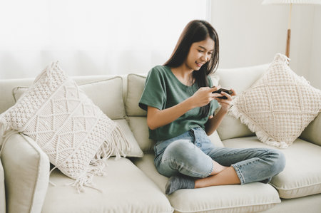 Happy young asian woman relaxing at home she is sitting on sofa and using mobile smartphone. She is playing online gaming have funの写真素材
