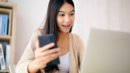 Young asian woman happy working on computer laptop and hand typing chat message on mobile smartphone at home. Video call, Online meetingの写真素材