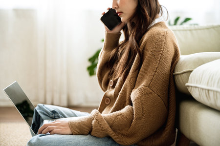 Asian woman using mobile smartphone and typing on keyboard computer laptop in living room at homeの写真素材