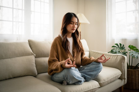 Young asian woman hand meditating and sitting on couch in living room at houseの写真素材