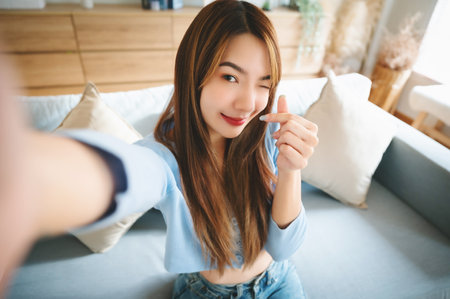 Pretty young asian female with big smile sitting at living room. She having fun taking light cheerful selfie on blurred backgroundの写真素材