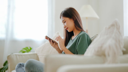 Happy young asian woman relax on comfortable couch at home texting messaging on smartphone, smiling girl use cellphone chatting, browse wireless internet on gadget, shopping online from homeの写真素材