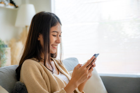 Happy young asian woman relax on comfortable couch at home texting messaging on smartphone, smiling girl use cellphone chatting, browse wireless internet on gadget, shopping online from homeの写真素材