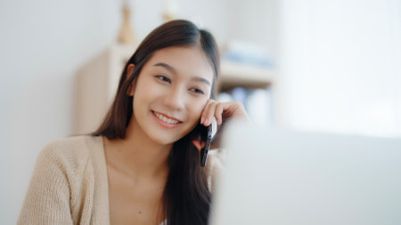 Young asian woman talking mobile smartphone while working on computer laptop on desk in living room at homeの写真素材