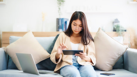 Young asian woman wearing glasses and using tablet while seated on couch at homeの写真素材