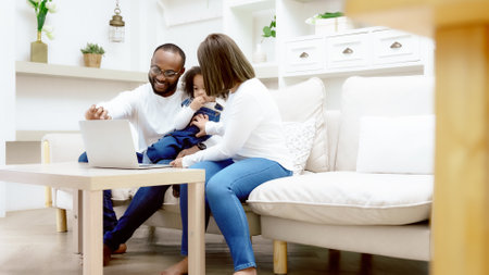 Happy african family parents and little children enjoy using computer laptop together sit on sofa, technology addicted couple with kids having fun at homeの写真素材