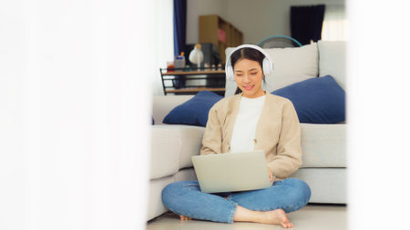 Young asian woman wearing headset in good spirits working on laptop at home while sitting on the floor close to the couchの写真素材