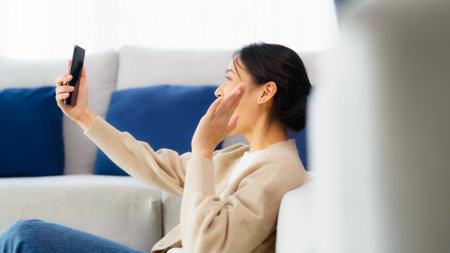 Happy young asian woman relaxing at home. Female smile sitting beside sofa and holding mobile smartphone. Girl using video call to friendの写真素材
