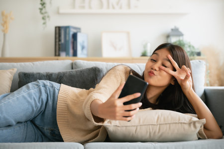 Happy young asian woman relax on comfortable couch at home texting messaging on smartphone, smiling girl use cellphone, browse wireless internet on gadget, shopping online from homeの写真素材