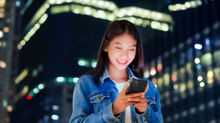 Beautiful young Asian woman walking in city street at night using mobile smartphone. Happy woman standing and typing messages on her mobile phoneの写真素材