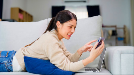 Young asian woman using smart phone at home. Smiling female making video call meeting smartphone at house, messaging or browsing social networks while relaxing on couchの写真素材
