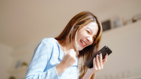 Funny euphoric young asian woman celebrating winning or getting ecommerce shopping offer on mobile smartphone. Excited happy girl winner looking smart phone celebrating successの写真素材