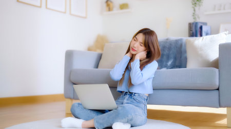 Office syndrome concept. Asian young woman working on laptop while seated on floor next to couch at home, feeling pain in neck and shoulder after working on computer laptop for a long timeの写真素材