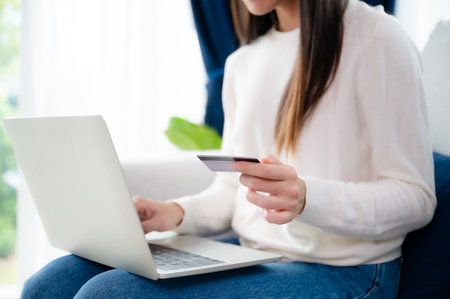 Young asian woman sitting on sofa in living room, makes online banking payments through the internet from bank card on computer laptop. Shopping online on notebook with credit cardの写真素材