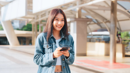 Young asian woman in jeans jacket clothes texting on smartphone walking on street in city on sunny dayの写真素材