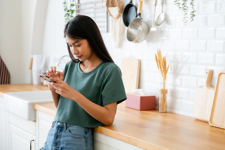 Happy young asian woman relaxing at home. Asia female standing at counter kitchen and using mobile smartphoneの写真素材
