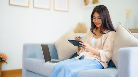 Young asian woman wearing glasses and using tablet while seated on couch at homeの写真素材