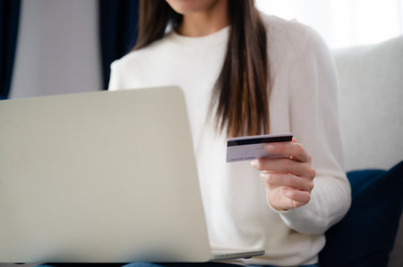 Young asian woman sitting on sofa in living room, makes online banking payments through the internet from bank card on computer laptop. Shopping online on notebook with credit cardの写真素材