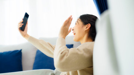 Happy young asian woman relaxing at home. Female smile sitting beside sofa and holding mobile smartphone. Girl using video call to friendの写真素材
