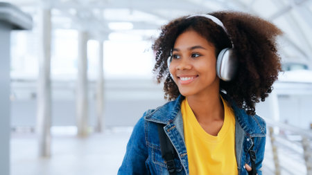 Happy young afro woman listening to playlist music with wireless headphones while wearing yellow shirt and jeans jacket outdoorの写真素材