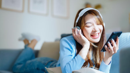 Happy young asian woman relaxing at home. Female smile sitting on sofa and holding mobile smartphone. Girl using video call to friend. Listening to musicの写真素材