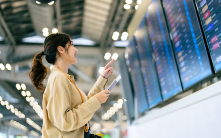 Young asian woman with passport and boarding pass as a hand in international airport looking at the flight information board, checking her flightの写真素材