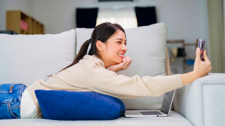 Young asian woman using smart phone at home. Smiling female making video call meeting smartphone at house, messaging or browsing social networks while relaxing on couchの写真素材