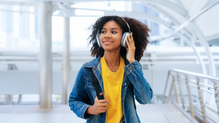 Happy young afro woman listening to playlist music with wireless headphones while wearing yellow shirt and jeans jacket outdoorの写真素材