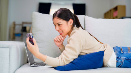 Young asian woman using smart phone at home. Smiling female making video call meeting smartphone at house, messaging or browsing social networks while relaxing on couchの写真素材