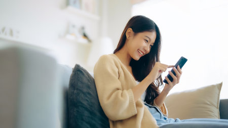 Happy young asian woman relax on comfortable couch at home texting messaging on smartphone, smiling girl use cellphone, chatting online message, shopping online from homeの写真素材