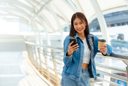 Young asian woman using mobile in city. Happy female tourist wearing jeans jacket and holding smartphone at publicの写真素材