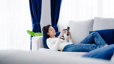 Young asian woman lying on sofa in living room, makes online banking payments through the internet from bank card on smartphone. Shopping online on mobile phone with credit cardの写真素材