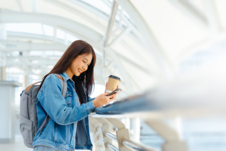 Young asian woman using mobile in city. Happy female tourist wearing jeans jacket and holding smartphone at publicの写真素材