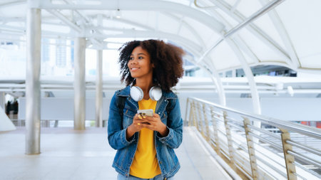 Happy young afro woman listening to playlist music with wireless headphones while wearing yellow shirt and jeans jacket outdoorの写真素材