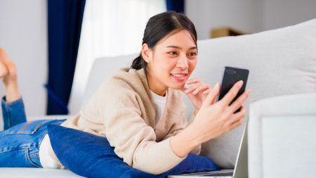 Young asian woman using smart phone at home. Smiling female making video call meeting smartphone at house, messaging or browsing social networks while relaxing on couchの写真素材
