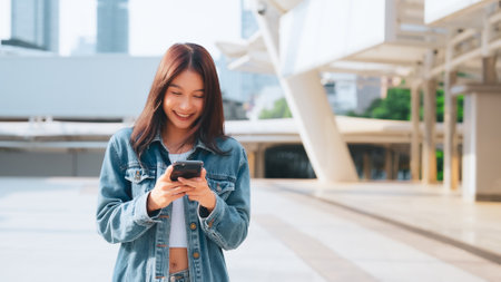 Young asian woman in jeans jacket clothes texting on smartphone walking on street in city on sunny dayの写真素材