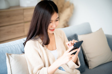 Happy young asian woman relax on comfortable couch at home texting messaging on smartphone, smiling girl use cellphone chatting, browse wireless internet on gadget, shopping online from homeの写真素材