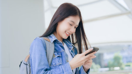 Young asian woman smiling using mobile smart phone outdoor. Happy female tourist wearing jeans jacket and holding smartphone at publicの写真素材