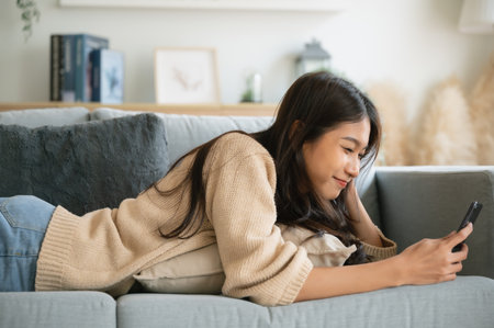 Happy young asian woman relax on comfortable couch at home texting messaging on smartphone, smiling girl use cellphone, browse wireless internet on gadget, shopping online from homeの写真素材