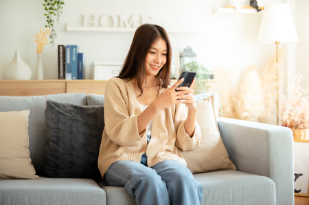 Happy young asian woman relax on comfortable couch at home texting messaging on smartphone, smiling girl use cellphone chatting, browse wireless internet on gadget, shopping online from homeの写真素材