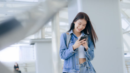 Young asian woman smiling using mobile smart phone outdoor. Happy female tourist wearing jeans jacket and holding smartphone at publicの写真素材