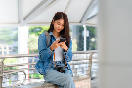 Young asian woman using mobile in city. Happy female tourist wearing jeans jacket and holding smartphone at publicの写真素材