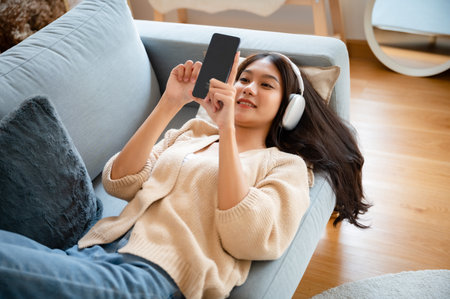Happy young asian woman relaxing at home. Female smile lying down on sofa and holding mobile smartphone. Girl using video call to friendの写真素材