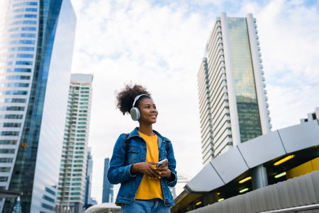 Cheerful black student girl wearing headset and using mobile smartphone, Walking at college building outdoor, Happy and smiling woman teenager. Modern educationの写真素材