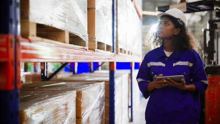 Young black woman warehouse workers holding digital tablet checking inventory management packaging boxes. Afro woman staff wearing vest and safety helmet and walking count the box at storehouseの写真素材