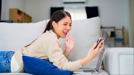 Young asian woman using smart phone at home. Smiling female making video call meeting smartphone at house, messaging or browsing social networks while relaxing on couchの写真素材