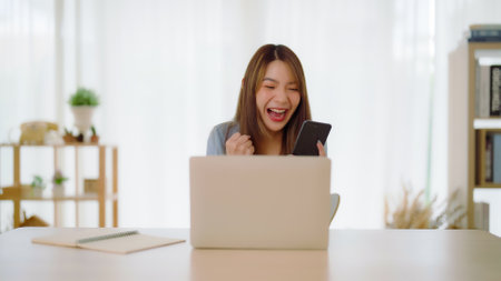 Funny euphoric young asian woman celebrating winning or getting ecommerce shopping offer on computer laptop. Excited happy girl winner looking at notebook celebrating successの写真素材