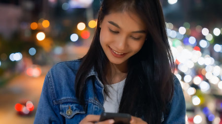 Vibrant urban nights, Young Asian woman, mobile chat in city. Technology, culture, and nightlife blend in modern lifestyleの写真素材