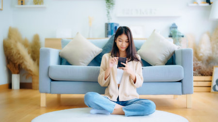 Happy young asian woman relax on comfortable couch at home texting messaging on smartphone, smiling girl use cellphone, chatting online message, shopping online from homeの写真素材